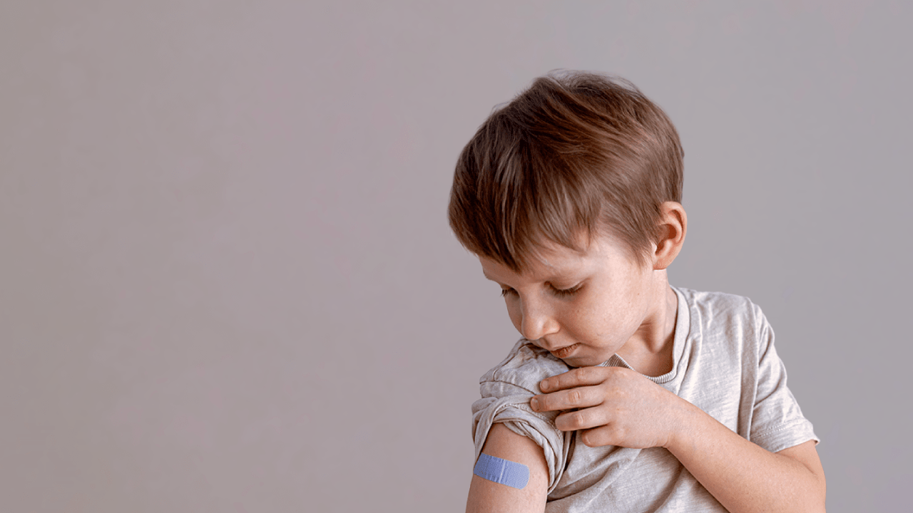 Don't risk it. Get vaccinated today A young boy lifts up the sleeve of his t-shirt and is looking at a blue band aid placed just below his shoulder, which shows where his flu vaccination was administered