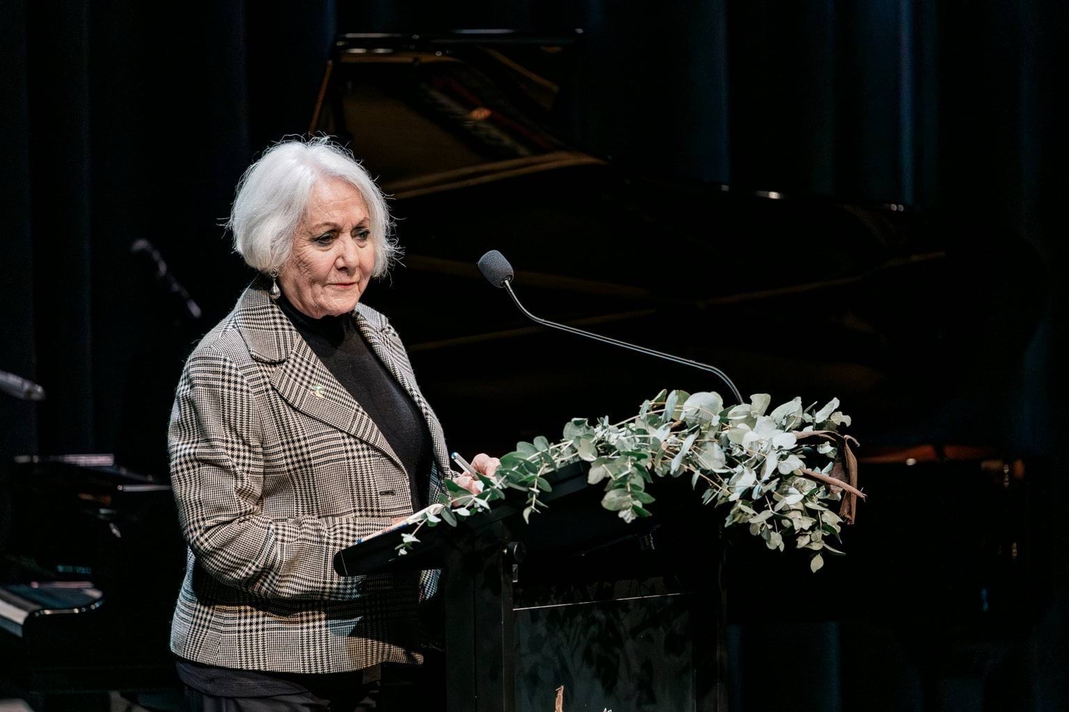 A lady with white hair and a grey jacket speaks at a podium with native flowers.