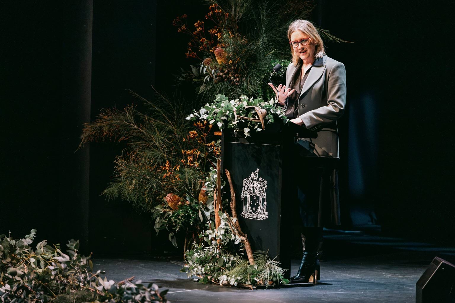 A person in a grey suit jacket and blonde hair speaks at a podium that is covered in flowers.