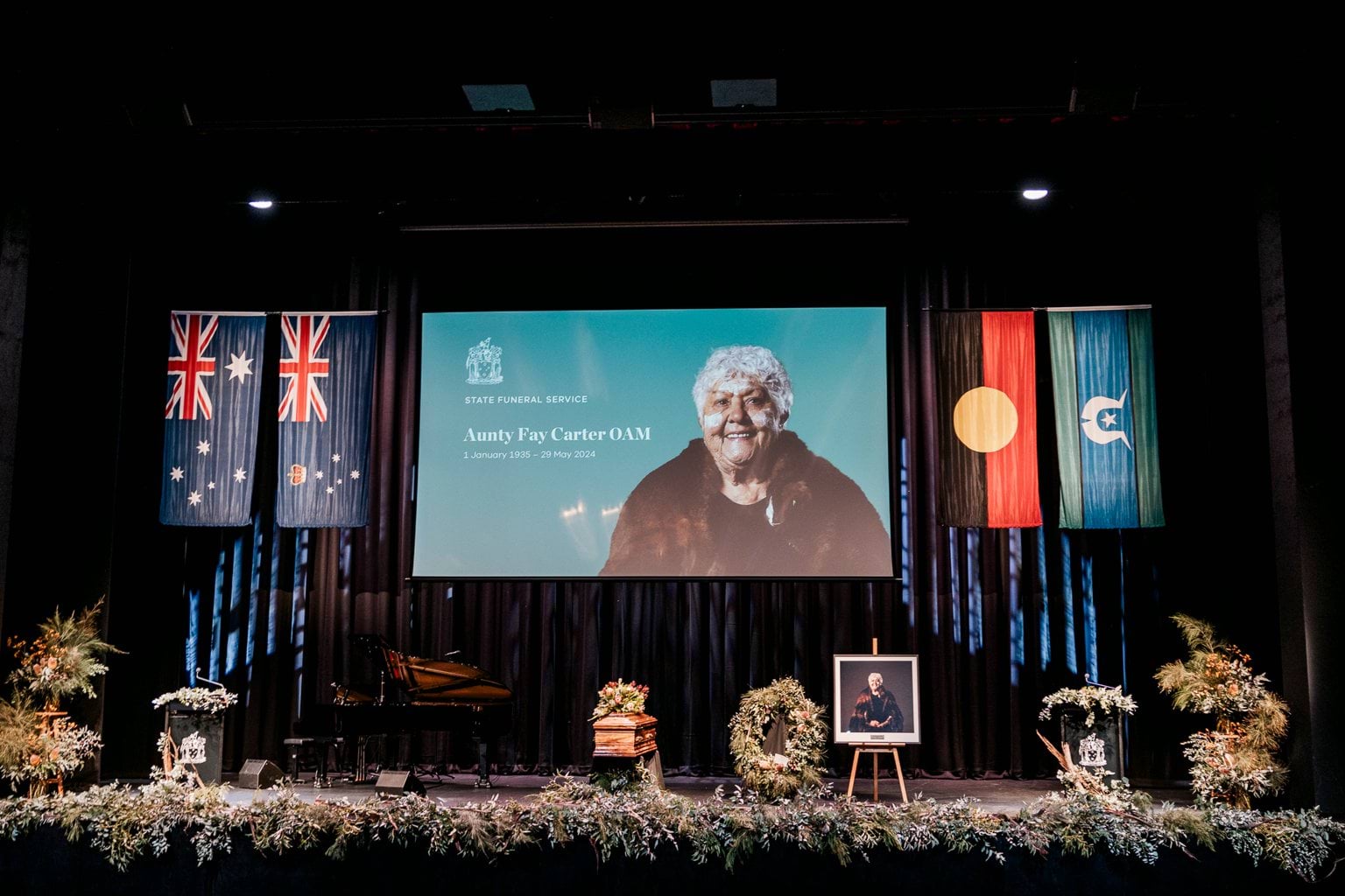 A stage with flowers, a coffin, four flags and a projection of an Aboriginal lady with the words 'Aunty Fay Carter OAM'