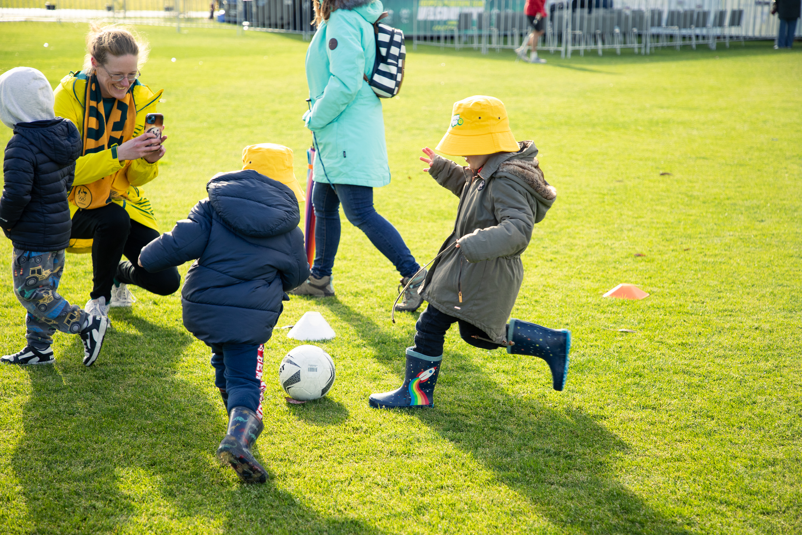 Children wearing yellow bucket hats and winter coats kicking a soccer ball on a field of green grass.