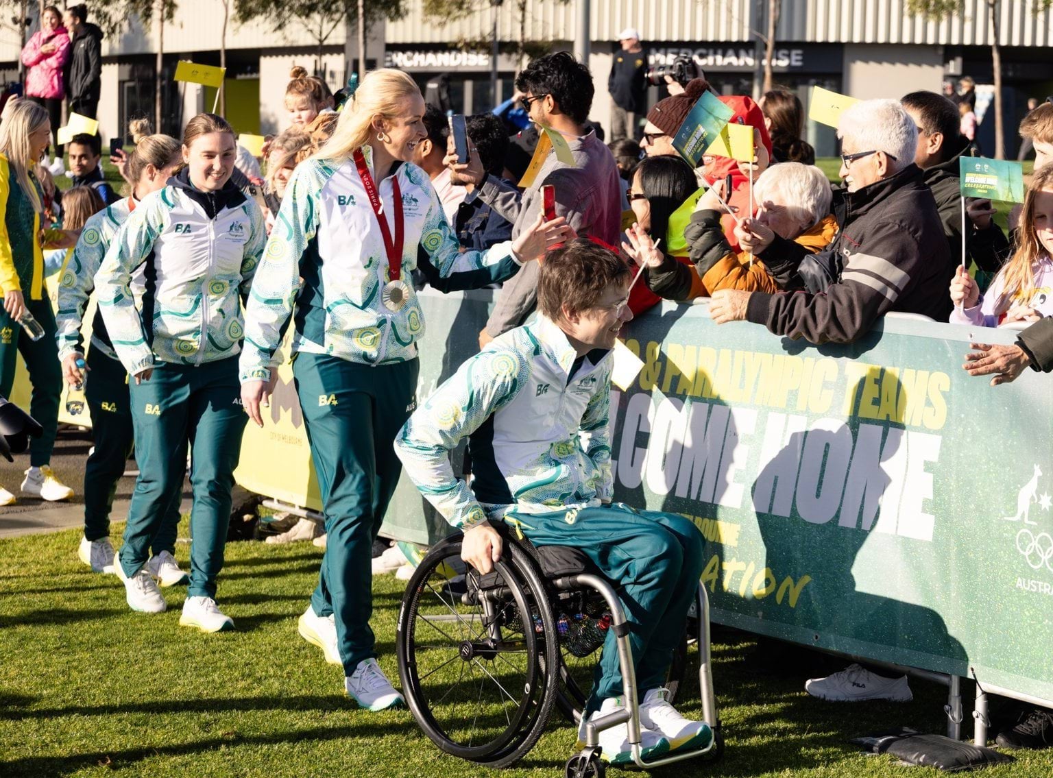 A crowd of people lining up behind a fence are waving flags as they greet a line of athletes wearing matching tracksuits, a person in a wheelchair leads the line of athletes with 3 others following behind on foot.
