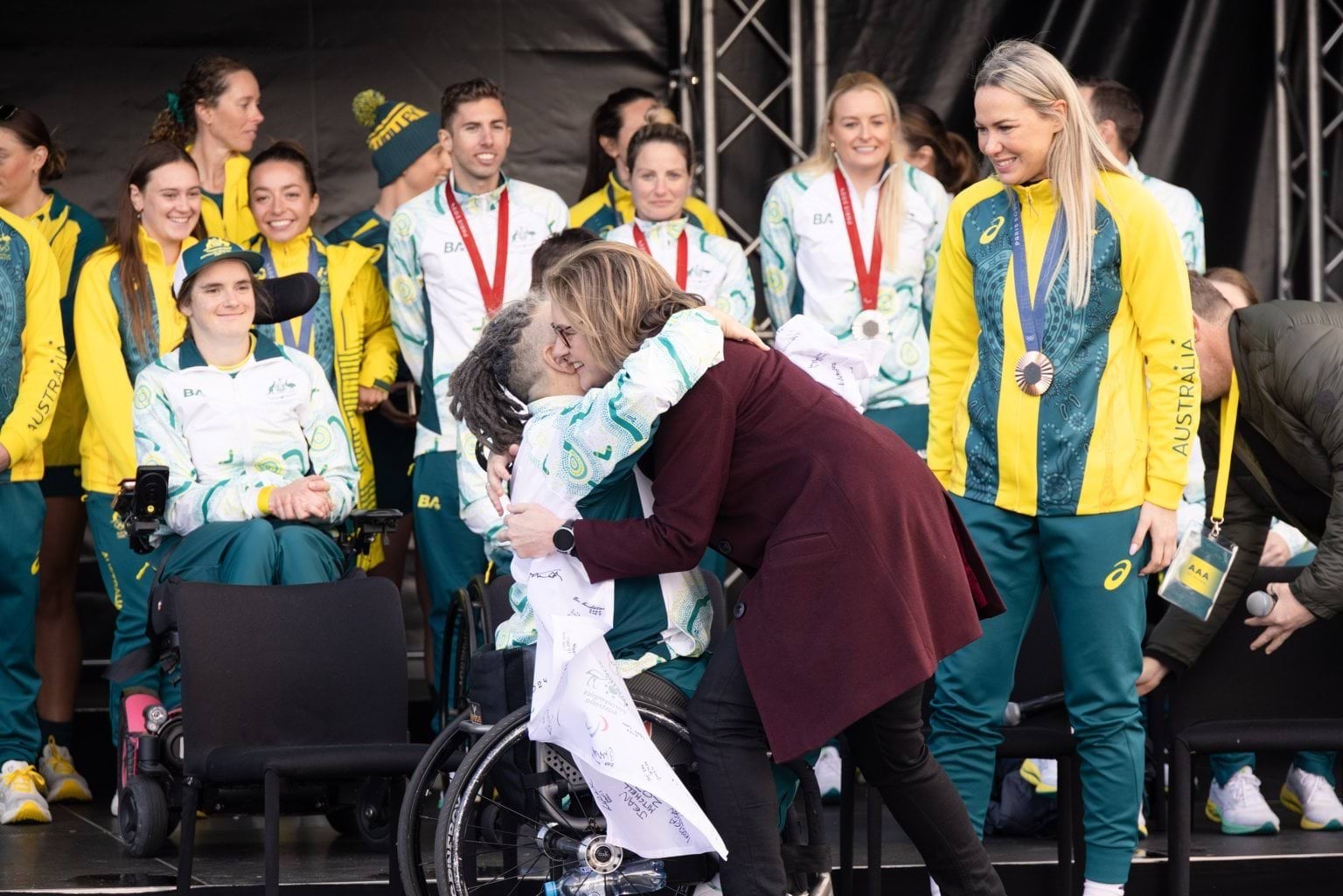 A woman with dark blonde hair in a long maroon coat hugs a person in a wheelchair.