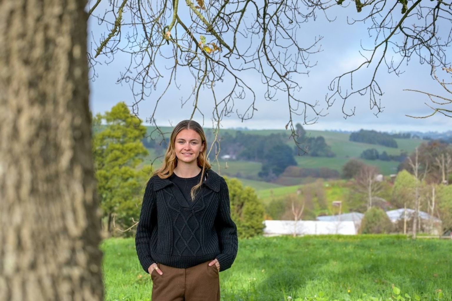 A woman standing on the grass with hills and trees in the background.