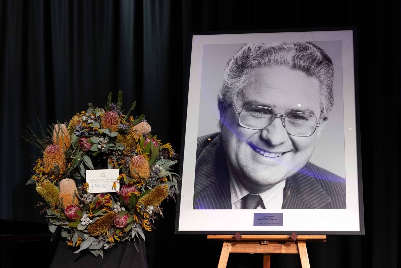 A wreath of native Australian flowers and a large framed black and white photo of a man wearing a suit with glasses.