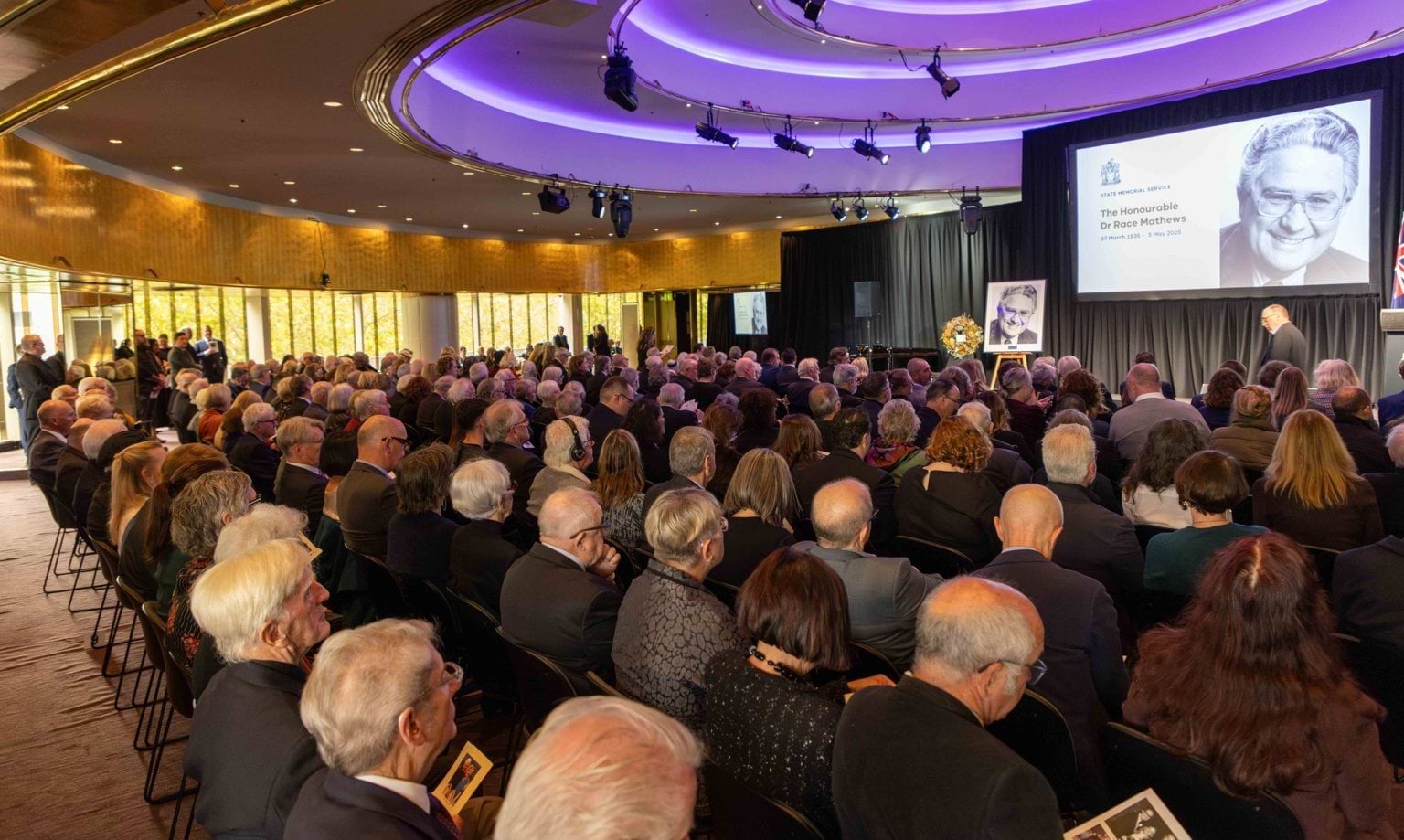 An audience sits in a round room watching a projection screen with a black and white photo of a man.