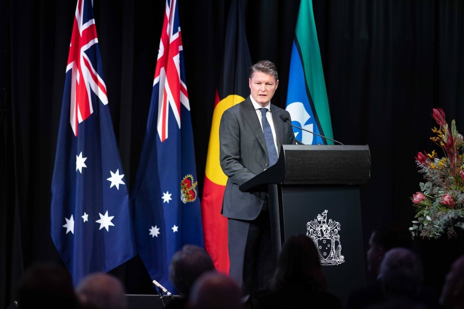 A man wearing a black suit and blue tie stands at a podium in front of flags.