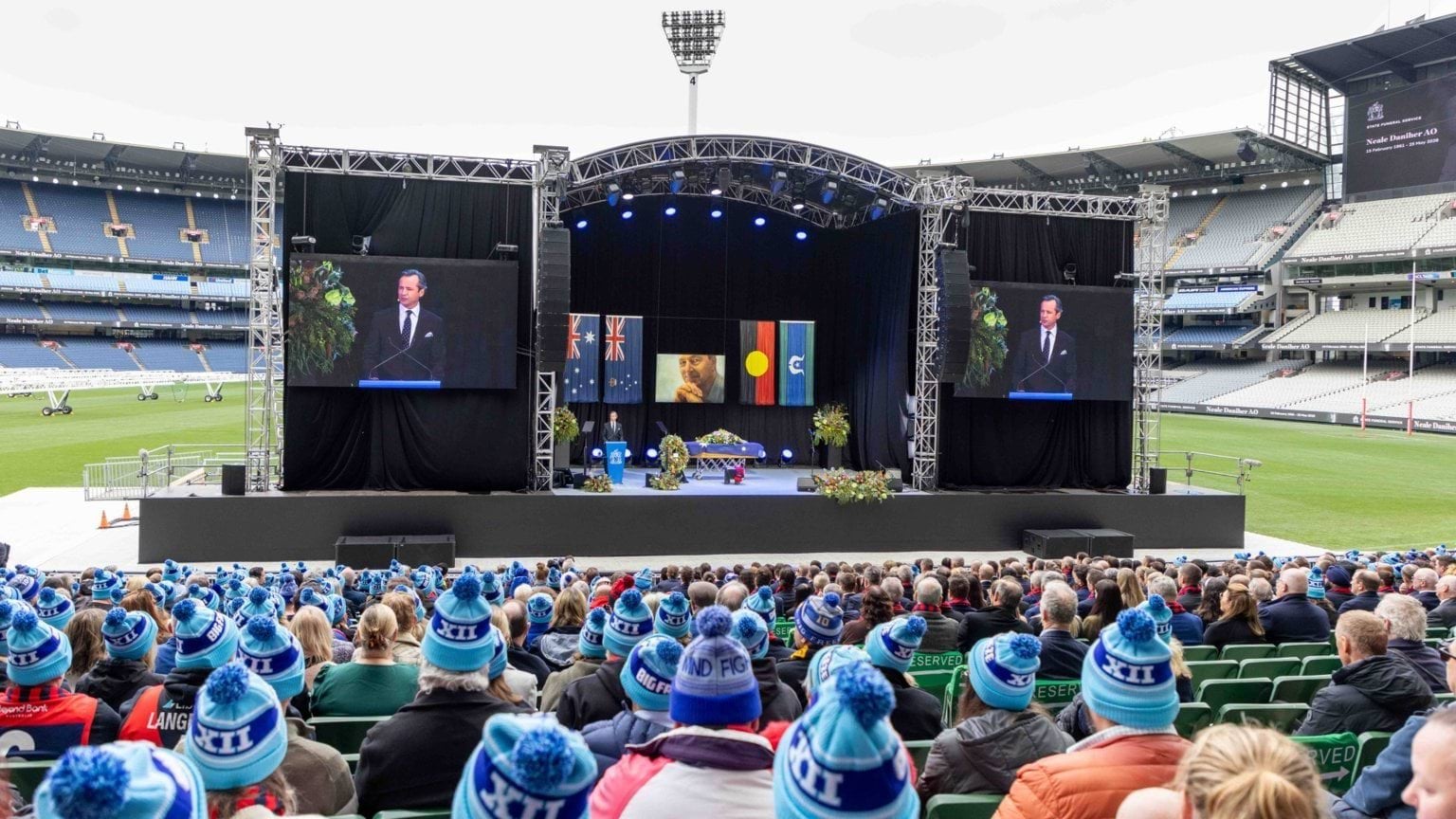 The image shows the stage for Neale Daniher AO's State Funeral Service, on the field of the Melbourne Cricket Ground, with audience members wearing FightMND beanies