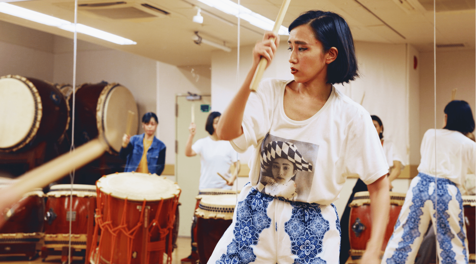 A person practices taiko drumming in a rehearsal studio, holding a drumstick above a drum while several other participants and large drums are visible in the background.