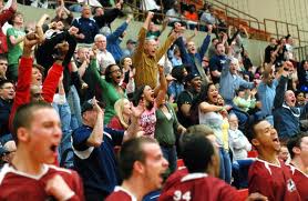 Photograph of a large, diverse group of cheering students, standing up and fist-pumping on the bleachers of a basketball game.