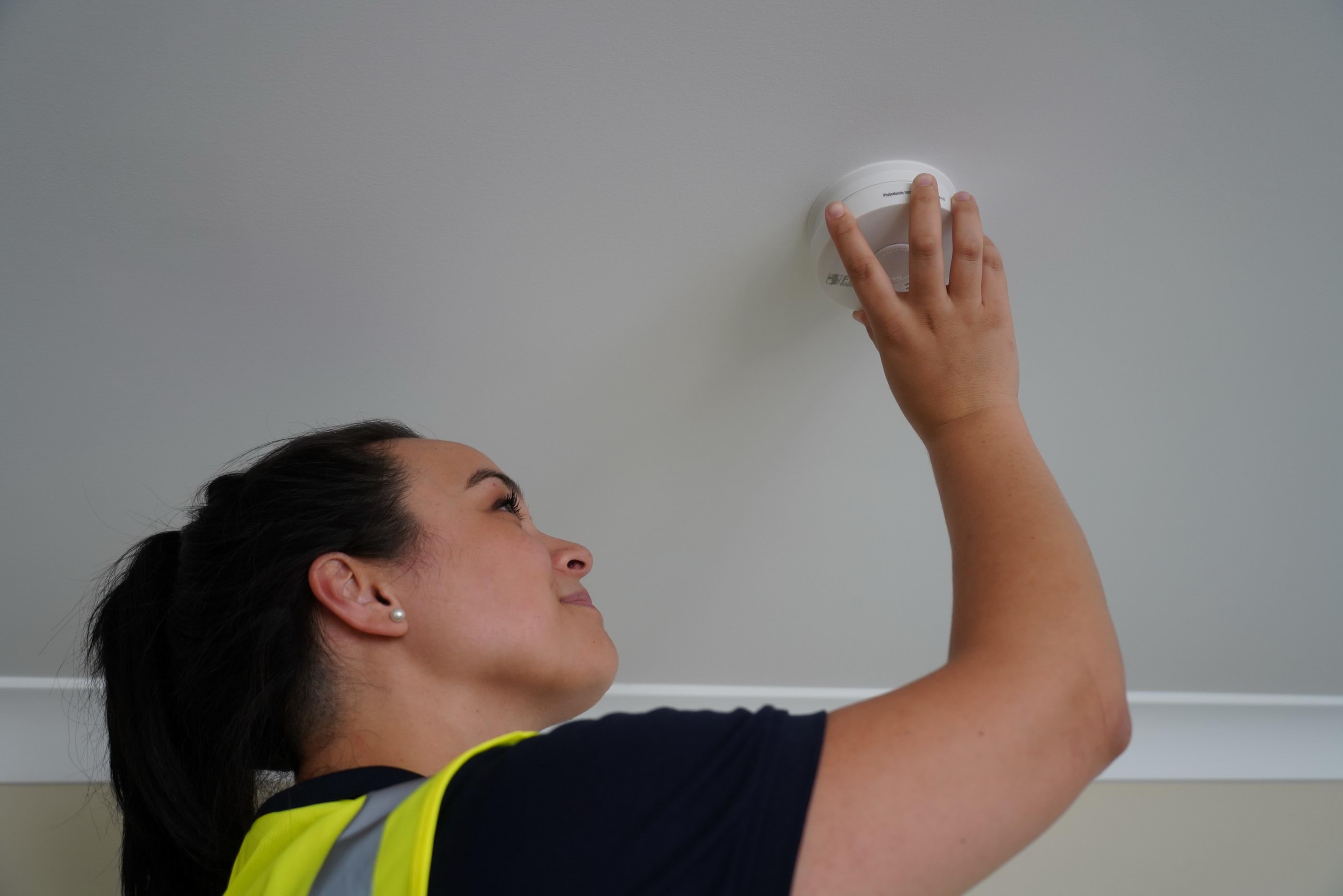 A woman in a high vis vest changes a smoke alarm attached to the ceiling