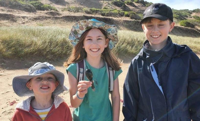 Owen, Lucy and Finn Dawson stand side by side on the beach at Ocean Grove on a sunny day. Sand dunes and vegetation can be seen in the background. Lucy, in the centre of the shot, is holding a satellite tag from an eel in her right hand.