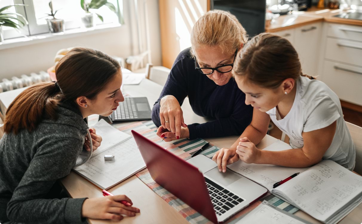 A mother home-schooling her two daughters at the kitchen table. There are schoolbooks and laptops scattered around.