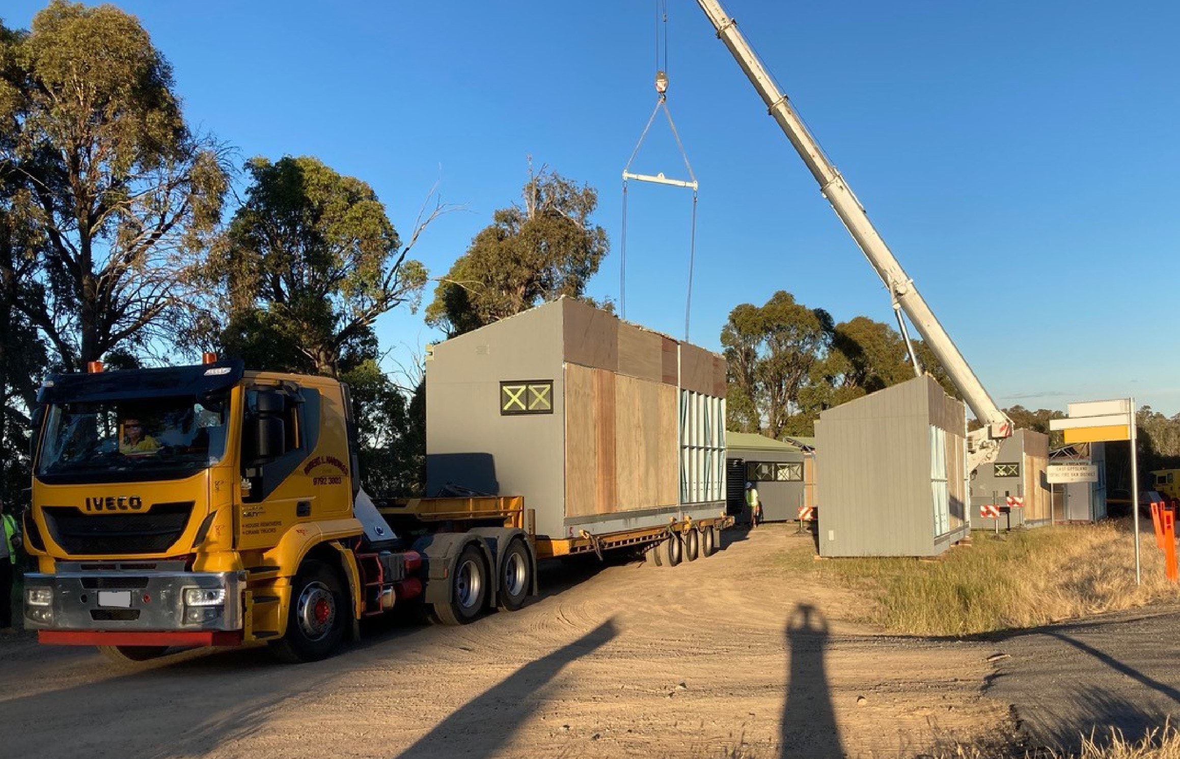 A truck loads a short-term housing module onto the back of the truck with the help of a crane