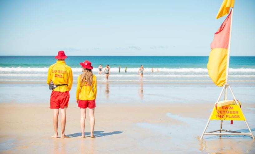 Two lifesavers at the beach, standing beside a flag with sign saying 