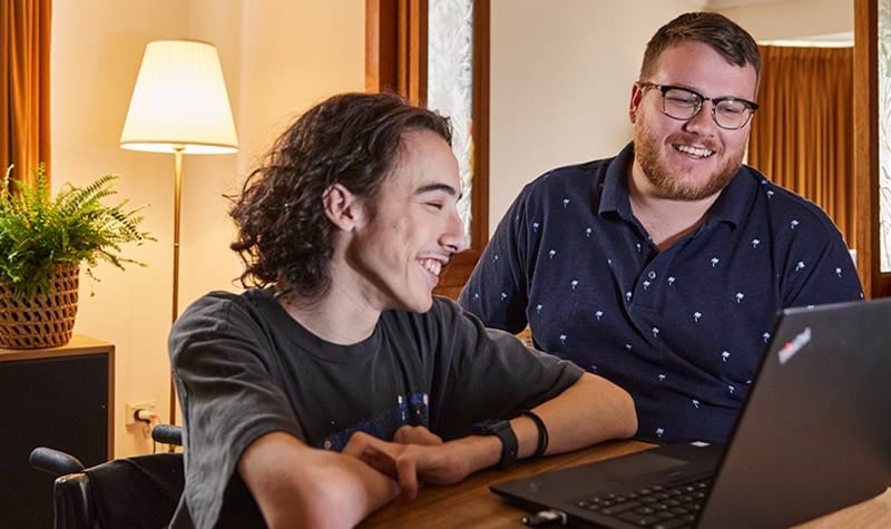 A client sitting at a desk with a disability worker and looking at a laptop