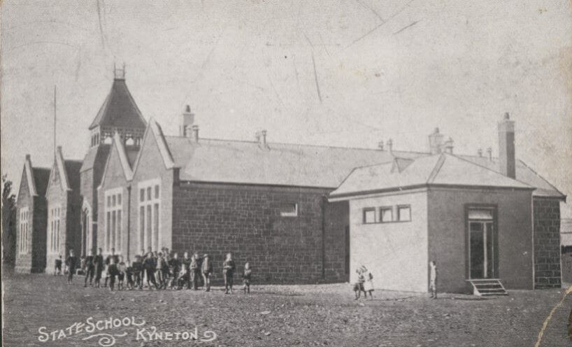 Black and white photograph of the Kyneton school building with children playing in the yard in front of the school. 