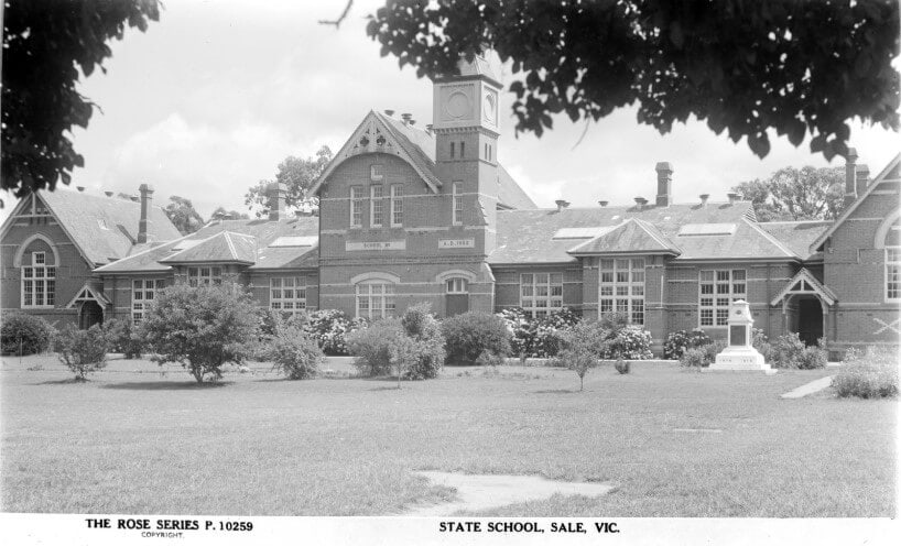 Black and white photograph of the Sale school building with trees and bushes on the grass in front of the school. The school building is brick and has a bell tower. 