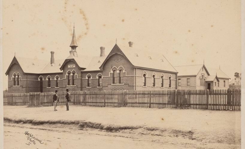 Black and white photograph of the Stawell school building. The brick building has a bell tower. Two men are standing in conversation in front of the school's picket fence. 