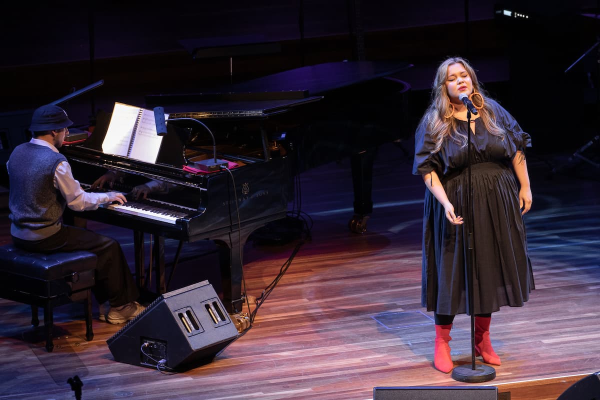 Bumpy stands at a microphone singing. Jake Amy is playing a grand piano on stage behind her. State Memorial Service for Dame Olivia Newton John AC DBE 