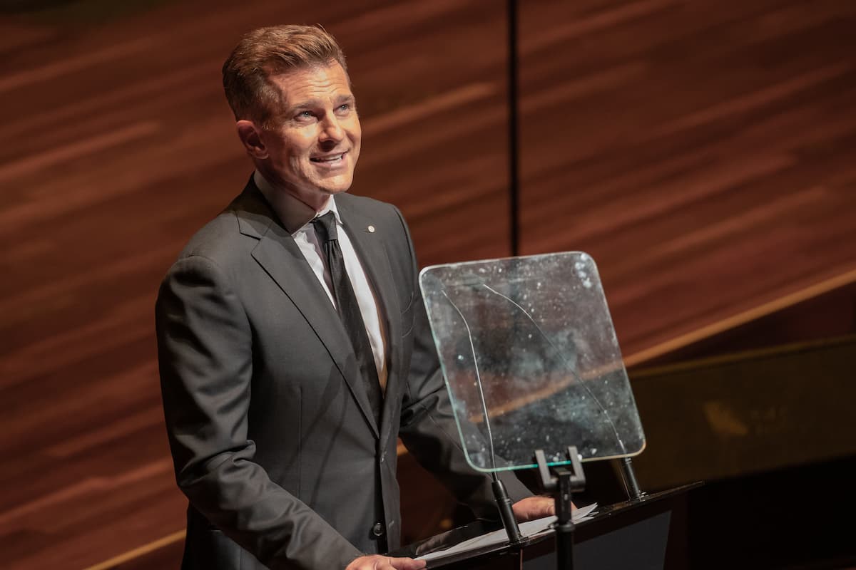 A man in a grey suit stands at a podium speaking to an audience