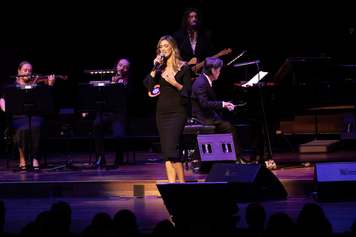 A woman in black sings on stage. People are playing instruments behind her.