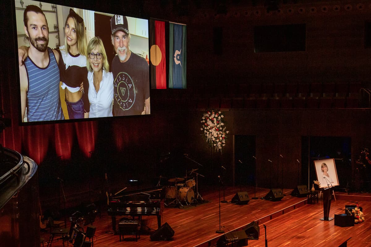 A portrait of a woman is on an empty stage with a family photo displaying on big screen in the background