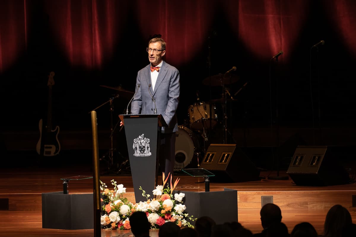 A man in a blue-grey suit with bowtie and glasses stands at a podium speaking to an audience