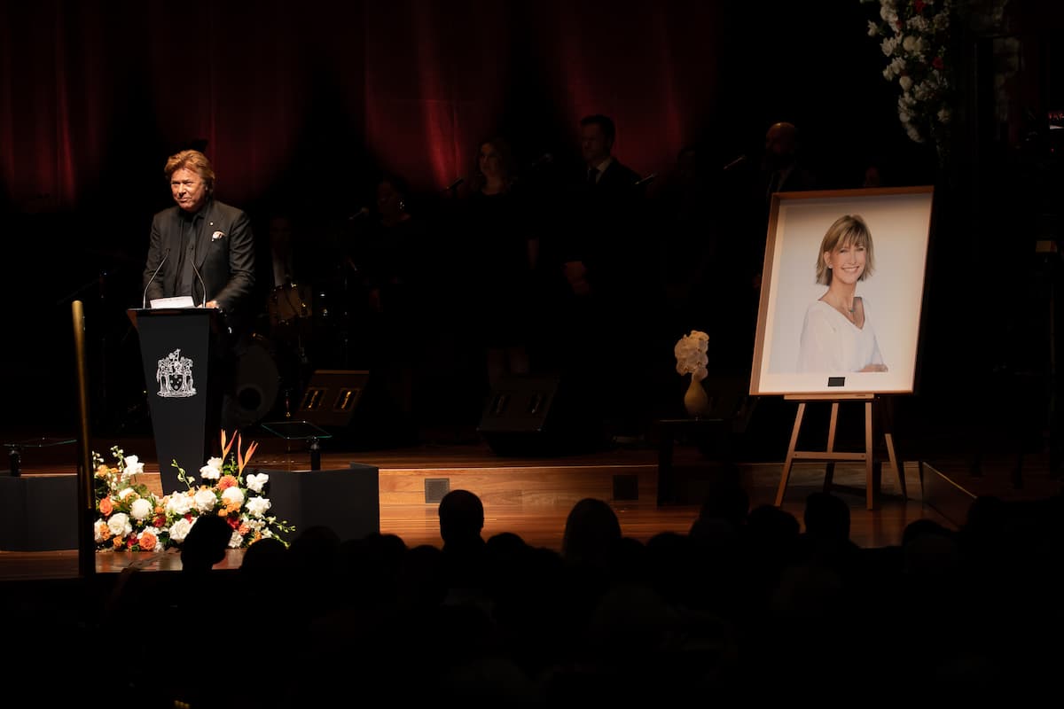 A man in a black suit speaks to an audience from a podium. To his right is a portrait of a smiling woman wearing all white.