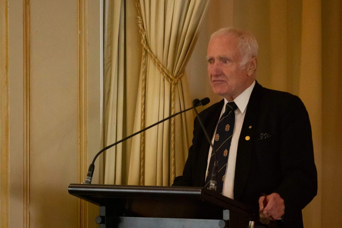 Robyn Williams speaks into microphones at a lecturn, behind him is a light coloured curtain