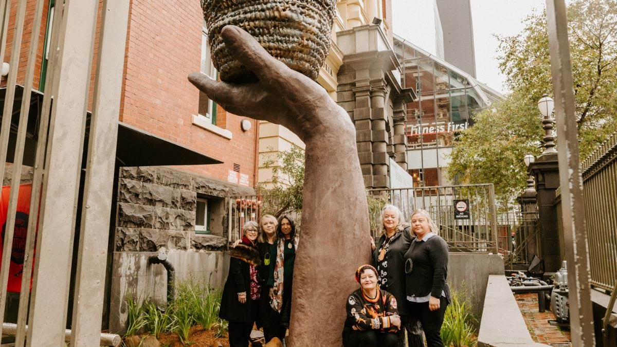 Creative Resilience artwork. A sculpture of a forearm and hand holding a basket. Located on Lonsdale Street outisde the Queen Victoria Womens Centre. Six member of the Ngardang Girri Kalat Mimini artists collective are gathered around the base of the scuplture.