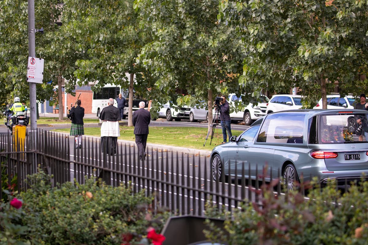 A bagpiper, priest and a funeral director walk down a tree-lined street as a grey hearse follows behind them.