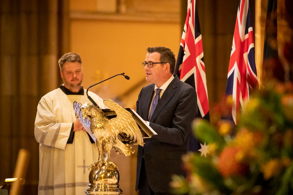 The Premier speaks into a microphone at a podium shaped like an Eagle. There are flags behind him.