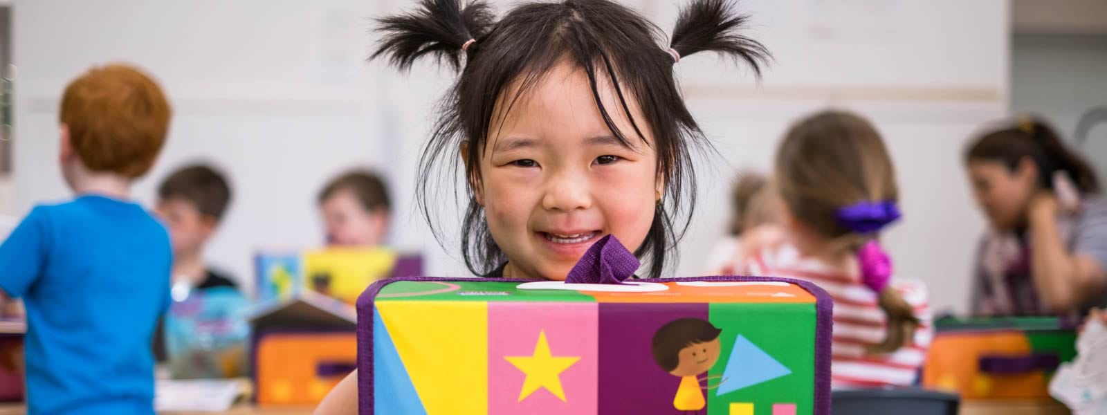 A young child with pigtails holds a colourful kinder kit, smiling. She's at kinder, with other children behind her opening their own kinder kits.