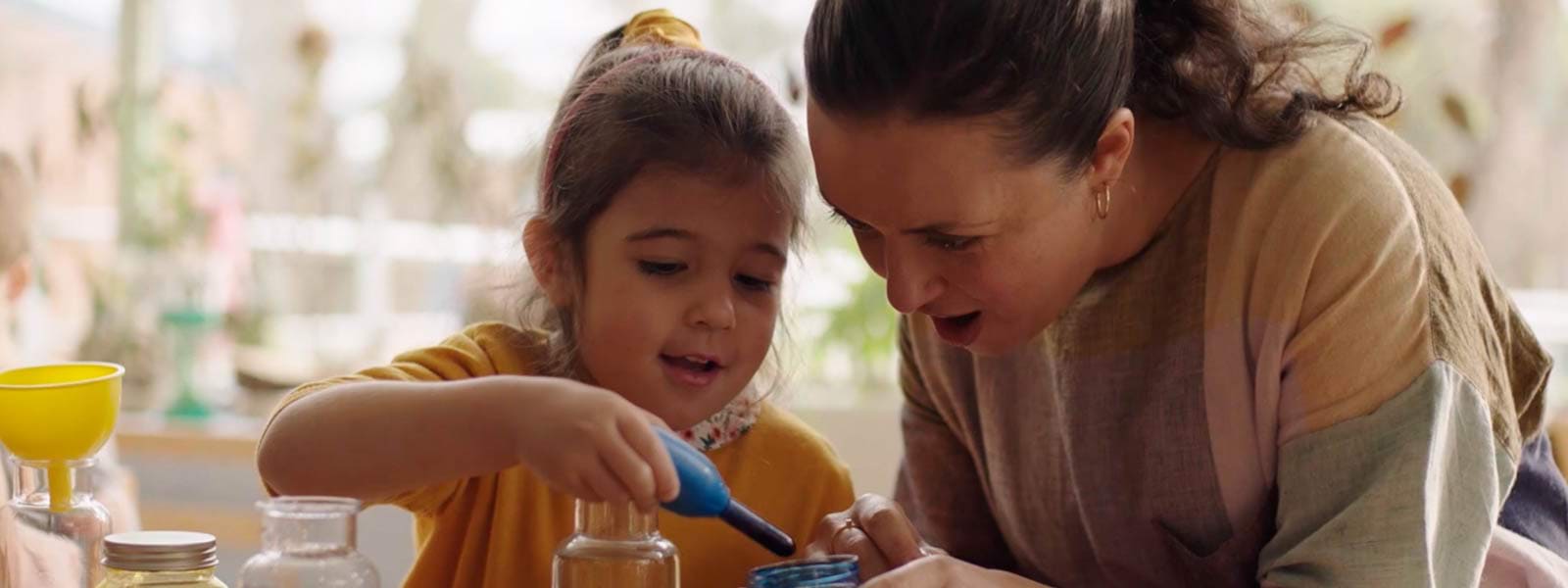 A woman helps a young child play with colourful dyes in a bright, sunny kindergarten