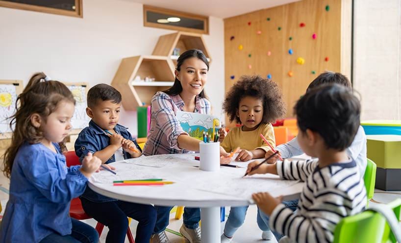 A teacher with a group of kids sitting around a table drawing and painting together.