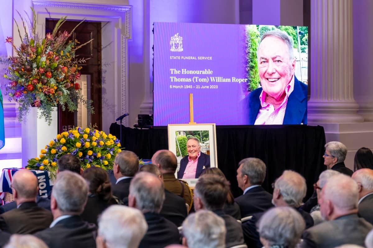 A room of people are seated and look towards a casket with flowers and pictures of a smiling man.