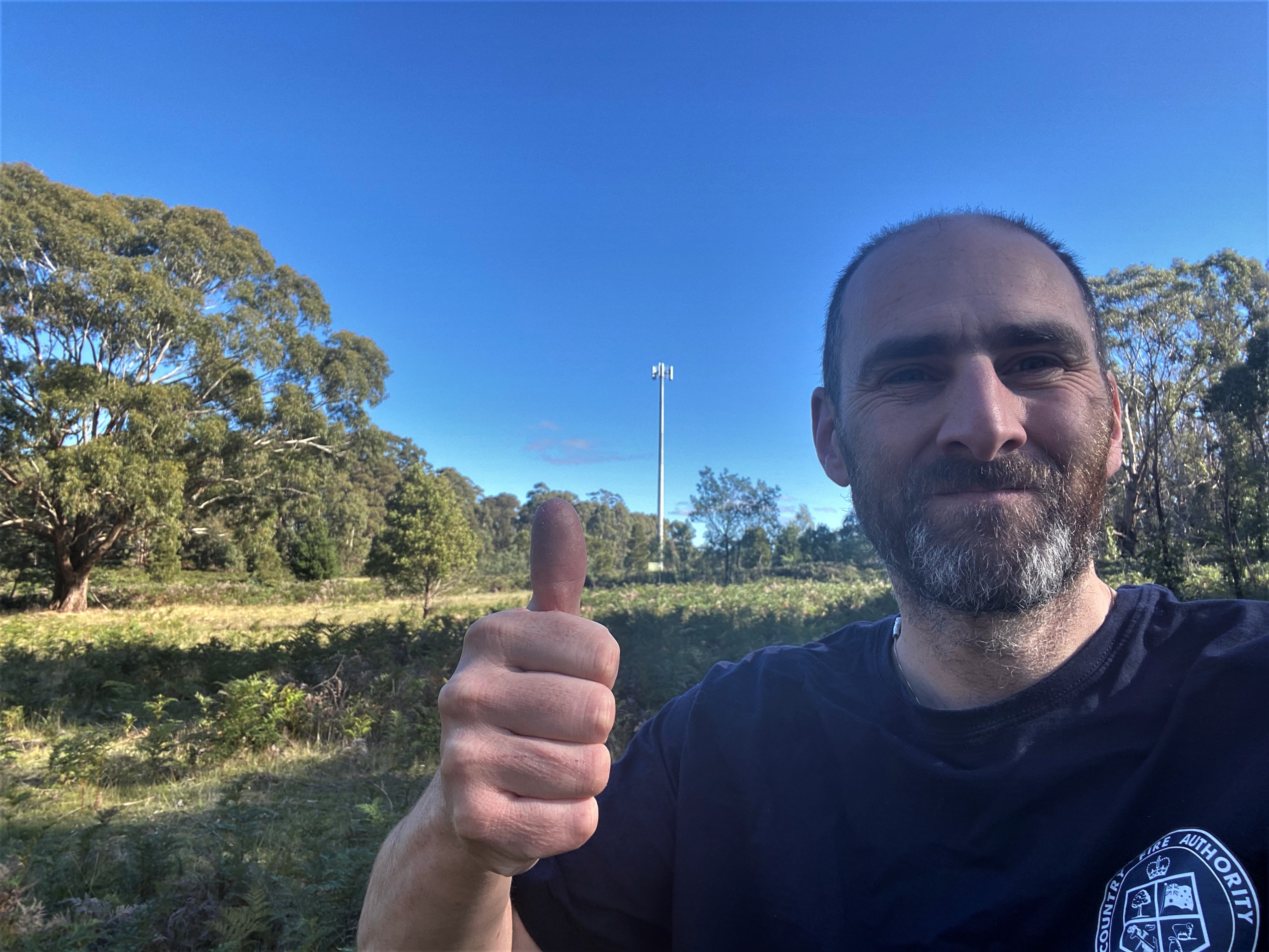 Man taking selfie on rural property with his thumb up and mobile phone tower in background