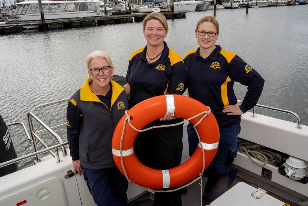 Image shows three people stood on a boat behind a life ring. 