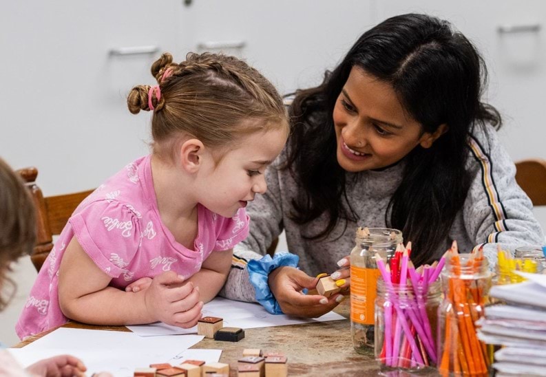 A woman and a young child smile and play with stamps, paper, and coloured pencils at a table.