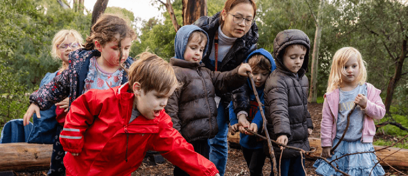 An educator and teachers play with sticks in puffer jackets outdoors.