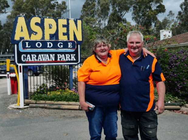 Image shows Yvonne and Russell Prince, owners of Aspen Lodge Caravan Park standing outside front of their business with caravan park sign