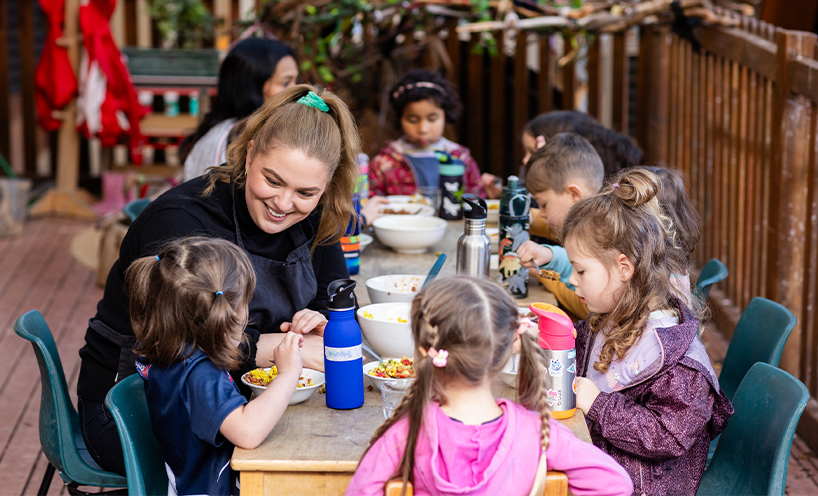 A kindergarten teacher sits with a large group of kindergarten children outside. The children are all eating lunch.