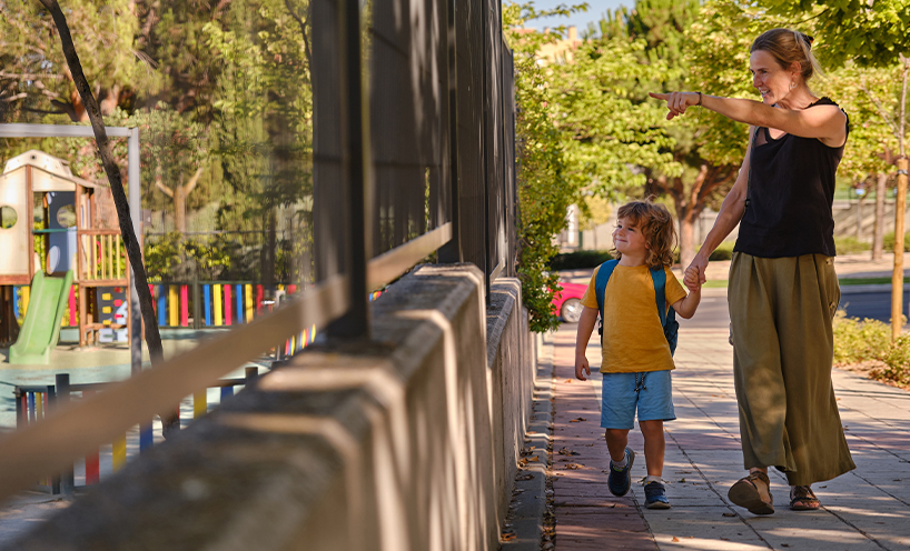 A mother and her child walk together holding hands. The mother is pointing off camera towards a playground.