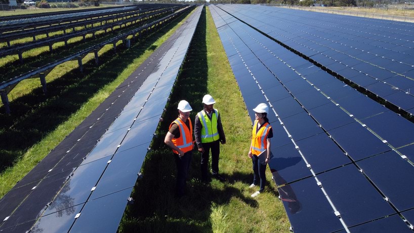 3 people standing in between long rows of solar panels in high vis vests and safety helmets in an outside field