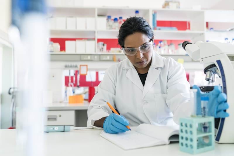 Woman working in a science lab with a white lab coat and safety glasses looking at a microscope sample