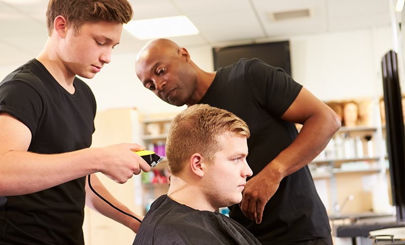 A teenage boy is buzz cutting a man's hair while his teacher assists him. All three of them wear black shirts.