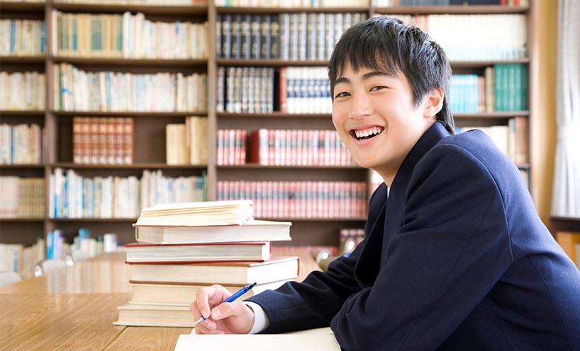 A high school student takes notes in their note book while smiling at the camera. Behind them is a stack of books and library shelves full of old books.