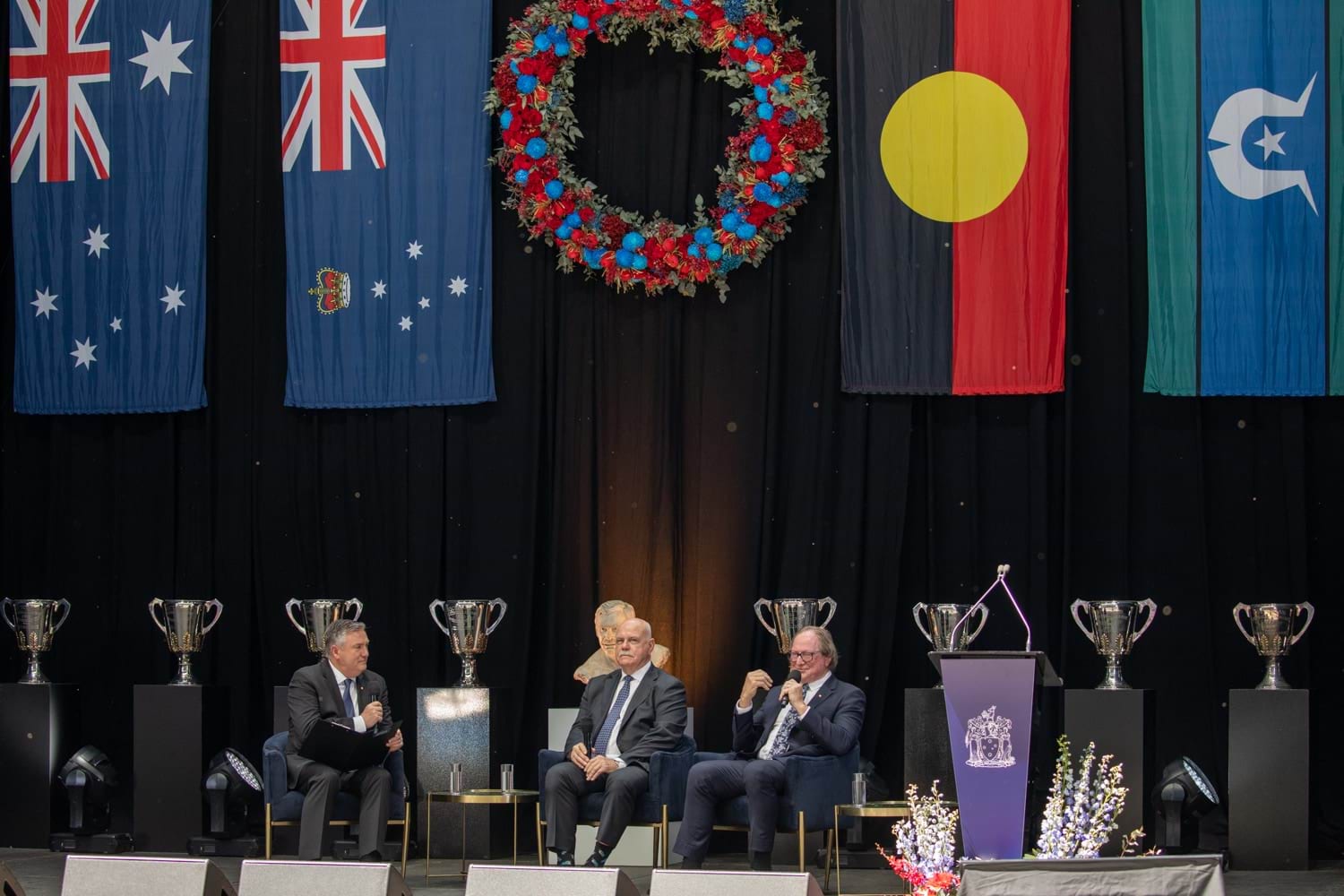 a stage is pictured that has 4 large flags draped in the background and is lined with AFL premiership cups in the background. There are three men in black suits sitting on chairs on the stage who are talking to each other and the audience through microphones 