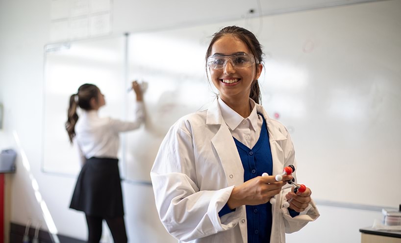 A teenage girl in a lab coat and protective glasses smiles to someone off camera. She is holding a DNA model in her hands.